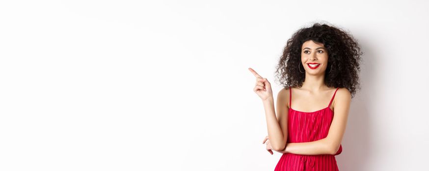 Elegant girl in red dress and evening makeup, pointing finger left and looking at logo, smiling satisfied, standing on white background.