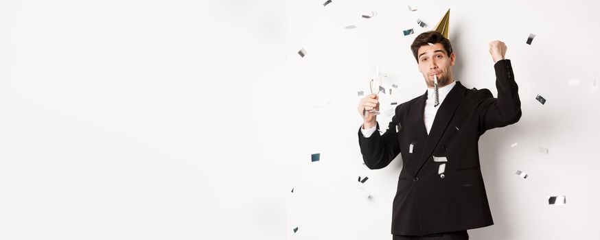 Handsome party guy in black suit having fun, celebrating new year, blowing whistle and drinking champagne while confetti falling, standing happy against white background.