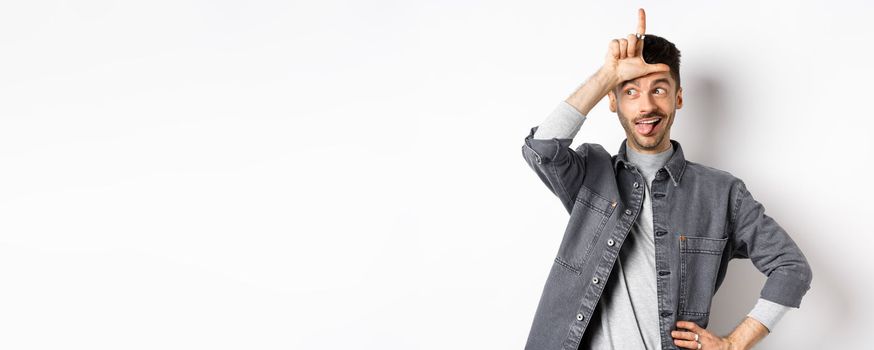 Funny guy mocking person, showing tongue and loser sign on forehead, making fun of lost team, standing on white background.