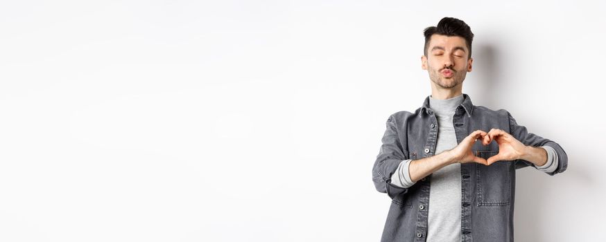 Cute young man waiting for kiss and showing heart gesture, confess on Valentines day, I love you sign, standing against white background.