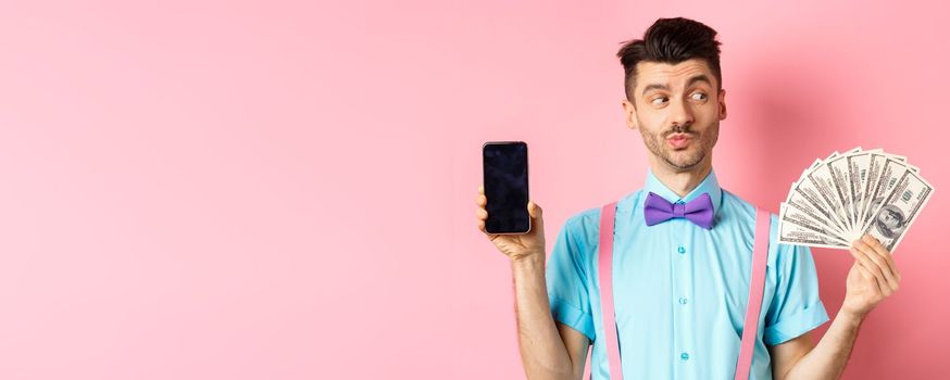 E-commerce and shopping concept. Thoughtful young man in bow-tie showing empty smartphone screen, looking pensive at money, standing on pink background.