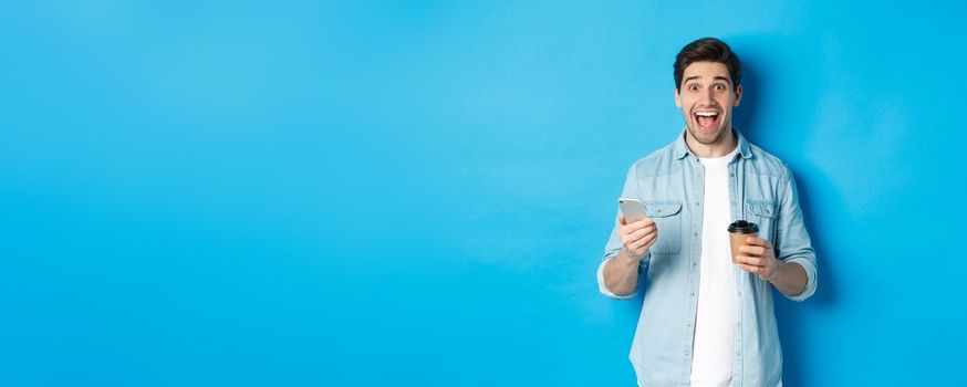 Happy young man drinking coffee and using mobile phone, looking excited, standing against blue background.