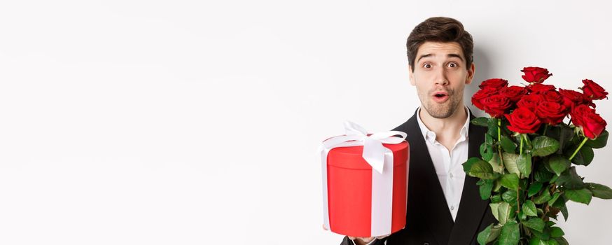 Close-up of attractive man in suit looking surprised, holding gift box and bouquet of roses, giving present for holiday, standing against white background.