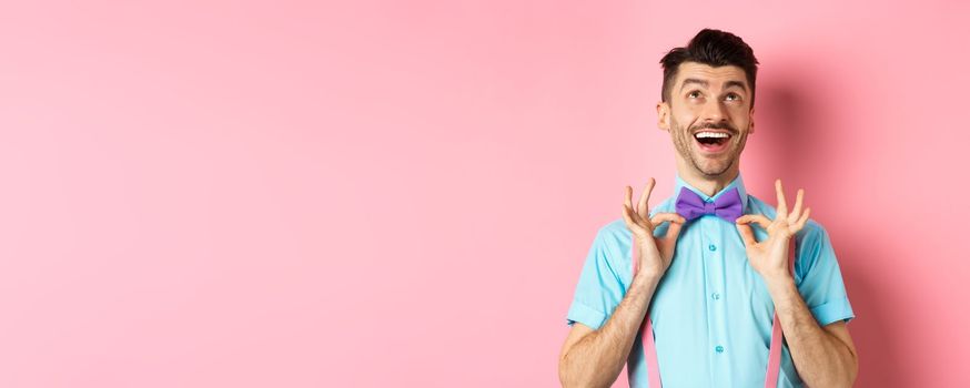 Happy young man with moustache, touching his bow-tie and laughing, looking up at logo, standing on pink background.