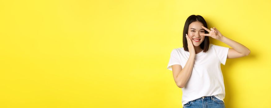 Lovely asian girl in white t-shirt posing with hand on cheek, showing peace sign on eye, standing over yellow background.