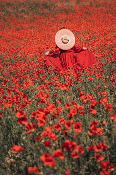 Young woman stands with her back in a long red dress and hat, posing on a large field of red poppies.