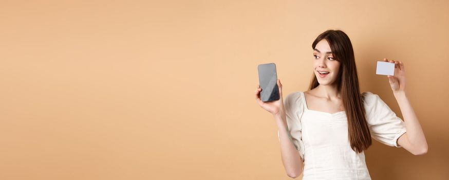 Check this out. Smiling happy young woman showing plastic credit card and empty mobile phone screen, demonstrate application, standing on beige background.