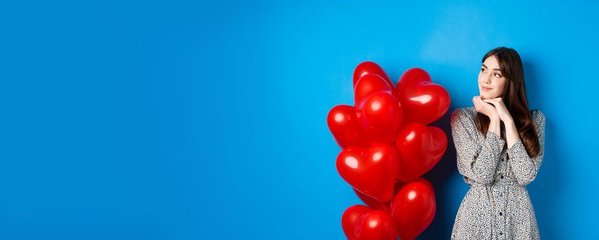 Valentines day. Dreamy beautiful woman in dress, imaging romantic date, standing near red hearts balloons and smiling, standing on blue background.