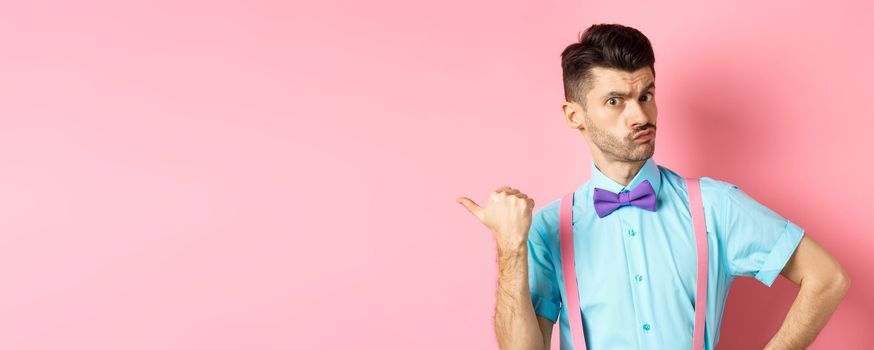 Serious-looking guy in suspenders suggessting to go outside, pointing left and looking confident at camera, starting fight, standing over pink background.