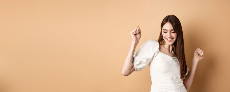 Carefree woman dancing and having fun, close eyes and smiling, standing on beige background.