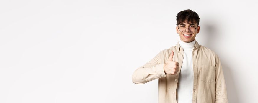 Positive smiling man showing thumbs up and looking satisfied, recommending and approving thing, standing pleased on white background.