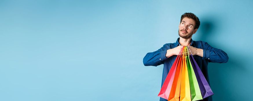 Dreamy young man holding colorful shopping bags and imaging something, looking at upper left corner empty space, blue background.