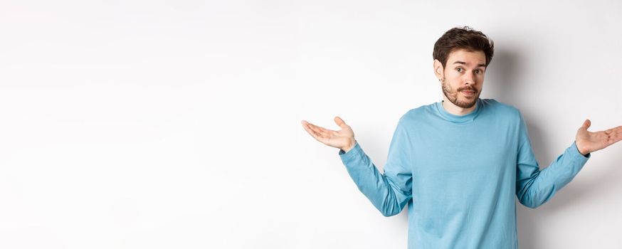 Clueless bearded guy in blue shirt, shrugging shoulders and looking unaware at camera, know nothing, standing on white background.