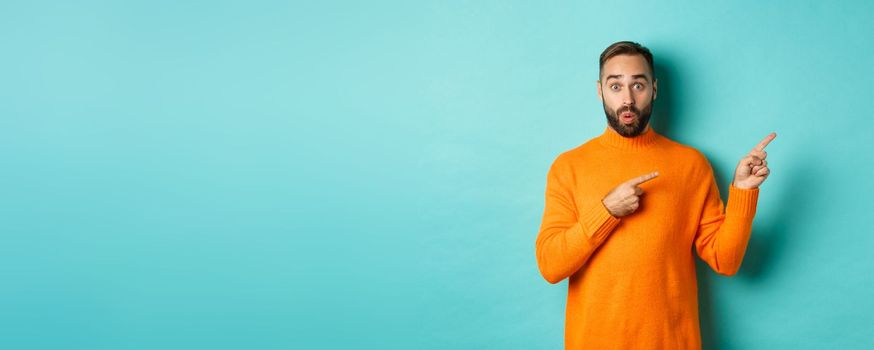 Amazed guy showing advertisement, pointing fingers right at banner, standing against turquoise background.