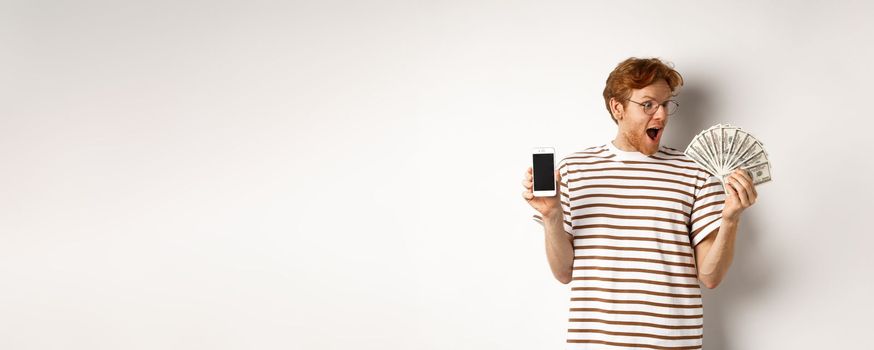 Amazed redhead man showing smartphone app on blank screen and money, winning prize cash online, standing over white background.