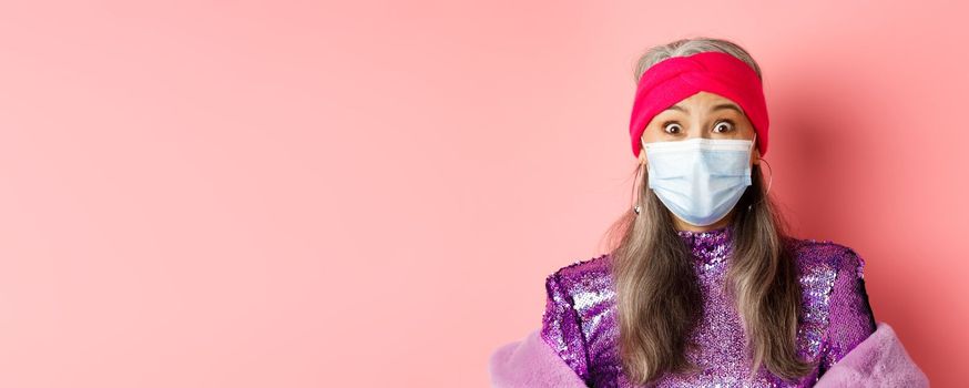 Covid-19, virus and social distancing concept. Close up of fashionable elderly woman in medical mask and headband staring surprised at camera, pink background.
