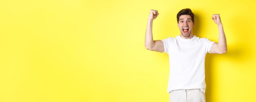 Image of excited man winning, raising hands up and celebrating, triumphing and rooting for team, standing over yellow background. Copy space