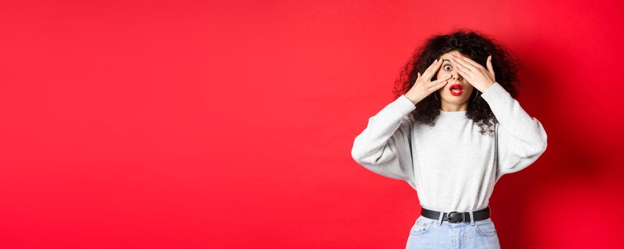 Shocked caucasian girl covering eyes and peek through fingers at something unbelievable, gasping speechless, standing on red background.