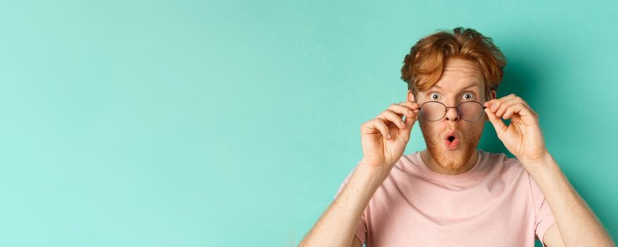 Surprised young man with red hair, checking out something cool, take-off glasses and saying wow impressed, standing over turquoise background.