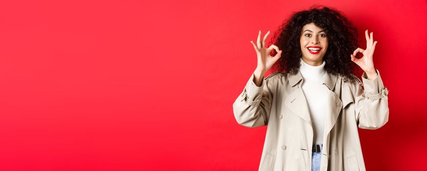 Stylish caucasian woman in trench coat showing okay gesture and smiling satisfied, recommending company, standing on red background.