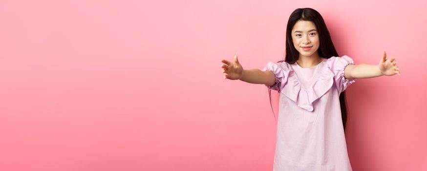 Cute teen asian girl stretch out hands for hug, want to cuddle, friendly greeting you, standing on pink background.