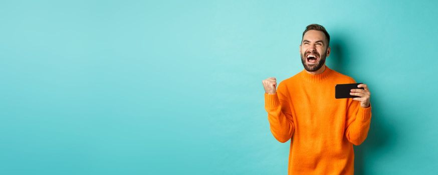 Image of man winning on mobile phone, making fist pump and rejoicing of victory, holding smartphone with satisfied face, turquoise background.