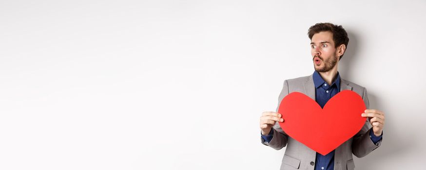 Man looking amazed at someone beautiful, holding big red heart cutout, standing on Valentines day over white background. Copy space