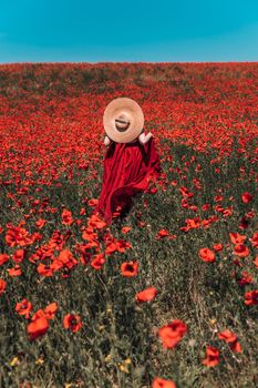 Young woman stands with her back in a long red dress and hat, posing on a large field of red poppies.