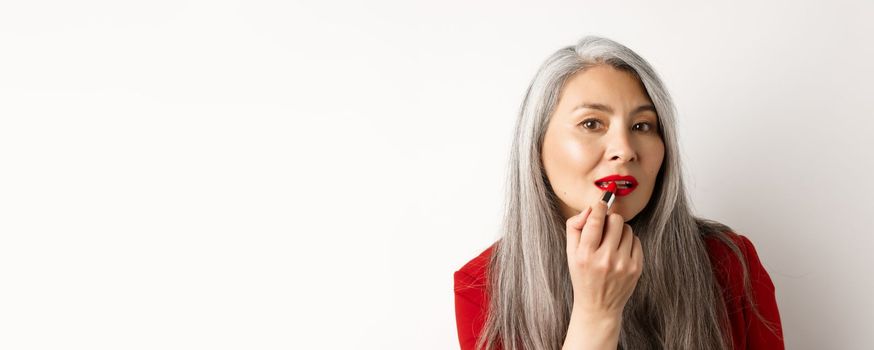 Beauty and makeup concept. Stylish asian mature woman with grey hair, looking in mirror and apply red lipstick, standing over white background.