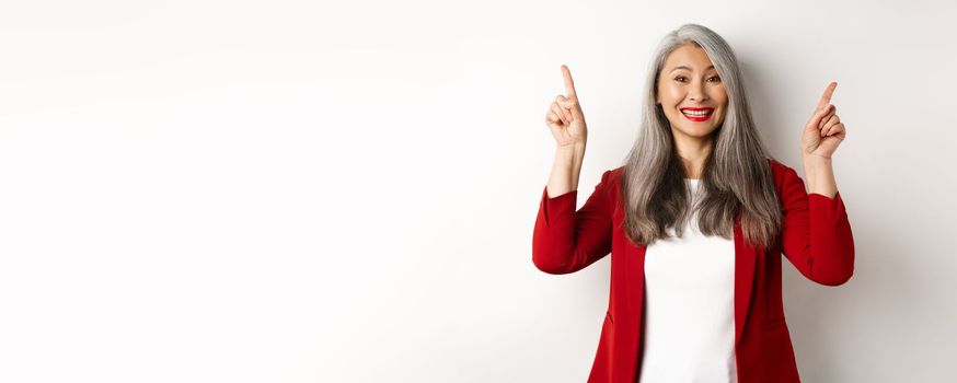 Happy mature woman in red blazer and makeup, smiling and showing advertisement on top, pointing fingers up at logo, white background.