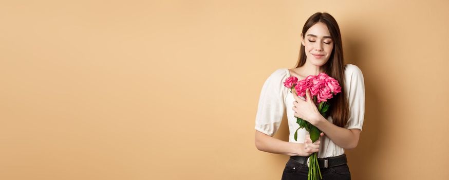 Valentines day. Tender and romantic girl, smell roses and smile with closed eyes. Girlfriend hugging gift flowers from lover, standing on beige background.
