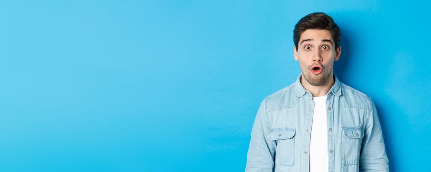 Close-up of handsome surprised man, open mouth and saying wow with amazement, hear announcement, standing over blue background.