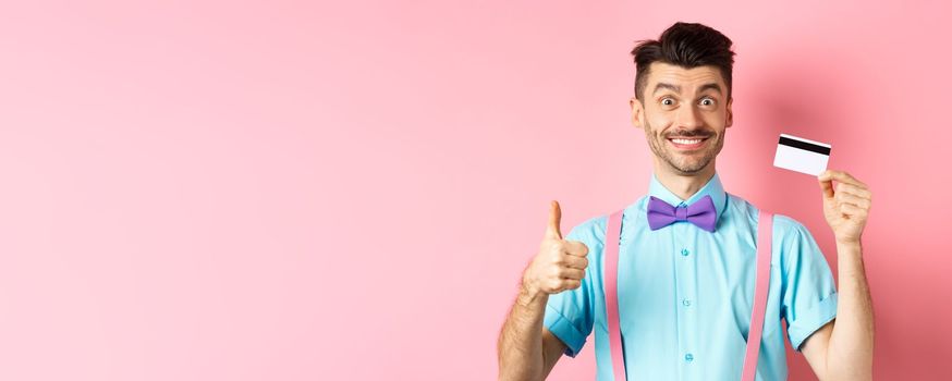 Cheerful guy in bow-tie showing thumb up and plastic credit card, like promo offer, smiling happy at camera, standing over pink background.