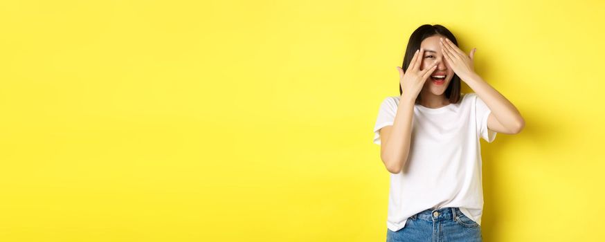 Intrigued asian girl waiting for surprise, peeking through fingers on eyes, smiling happy, standing over yellow background.