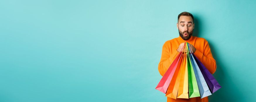 Excited man looking at shopping bags with purchases, standing amused against turquoise background. Copy space