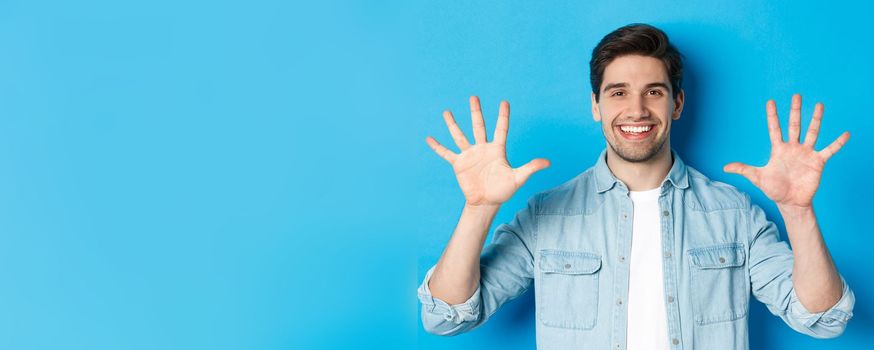 Close-up of handsome man smiling, showing fingers number ten, standing over blue background.