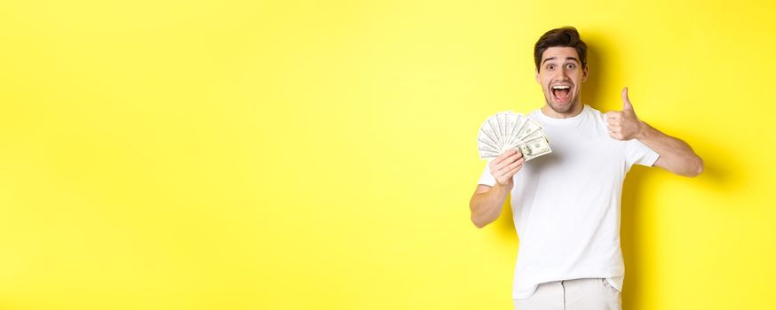Excited man holding money, showing thumb-up in approval, got credit or loan, standing over yellow background.