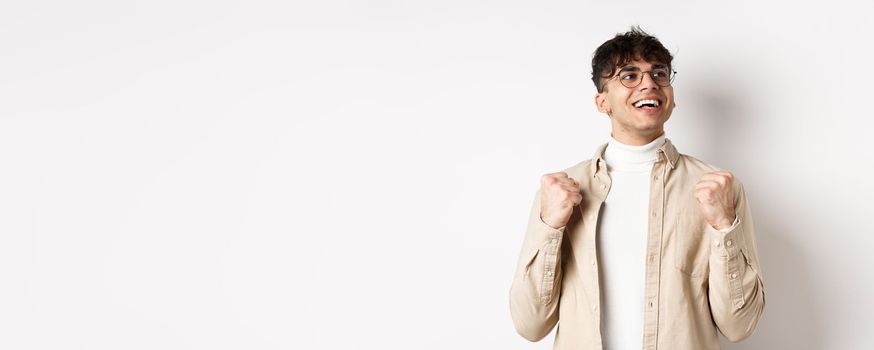 Lucky young man say yes with relieved face, smiling and making fist pump, triumphing of success, winning prize, standing on white background after achieving goal.