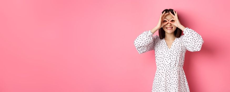 Carefree young asian woman looking through hand binoculars at camera, staring at discounts, standing over pink background.