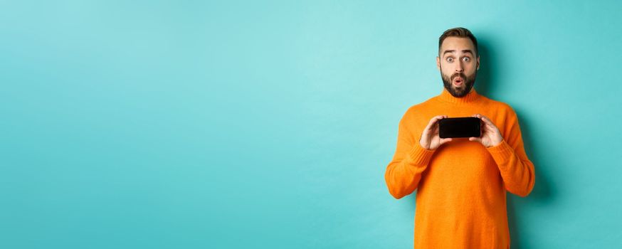 Impressed man showing smartphone screen, stare at camera amazed, demonstrate display, standing over light blue background.