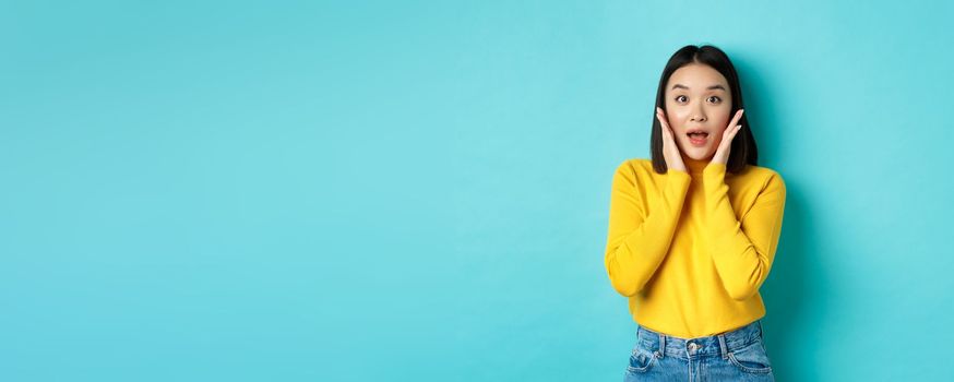 Portrait of surprised asian girl checking out promo, gasping amazed and touching cheeks, staring at camera amazed, blue background.