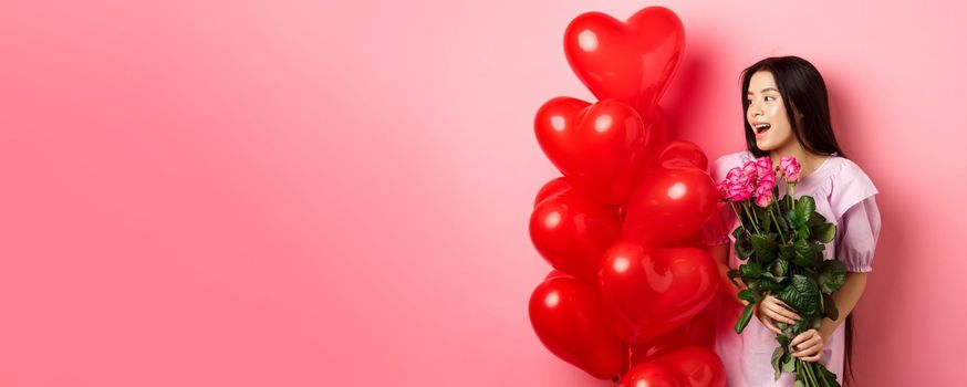 Portrait of asian teenage girl in love, holding flowers and looking at valentines day heart balloons, being on romantic date, pink background.