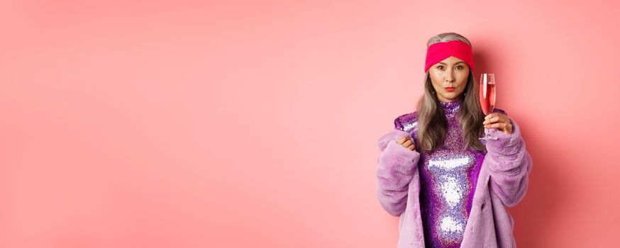 Fashionable asian senior female model raising glass of champagne, wearing trendy glitter dress and faux fur and looking at camera, congratulating with holiday, pink background.