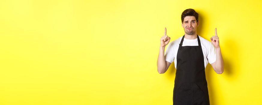 Displeased and skeptical waiter in black apron pointing fingers up, express dislike and indifference, standing over yellow background.