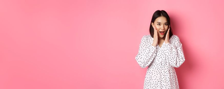 Young asian woman looking intrigued, gossiping and saying wow with curious face, looking interested at camera, standing over pink background.