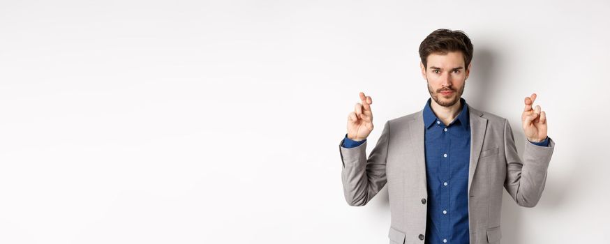 Hopeful man in suit cross fingers for good luck and looking confident in win, feeling determined, standing on white background.