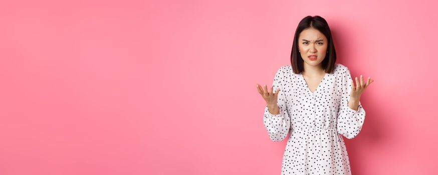 Angry and confused asian woman pointing hands at camera and grimacing furious, standing annoyed against pink background.