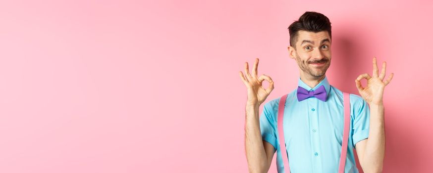 Portrait of handsome man with moustache and bow-tie, showing okay gestures and smiling in approval, recommending good offer, standing over pink background.