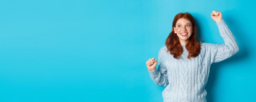 Happy redhead girl rooting for team, cheering with raised hand and smiling, celebrating victory or sucess, standing against blue background.