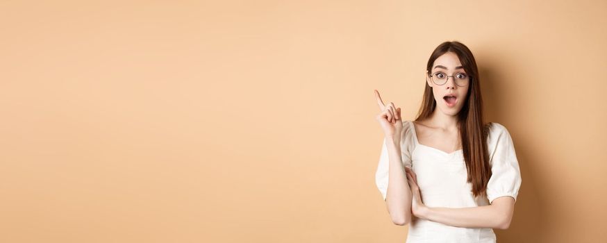 Creative young woman in glasses pitching idea, raising finger in eureka sign and gasping, have a plan, standing on beige background.